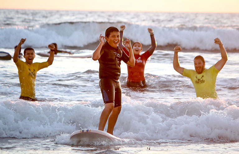 Photo of a camper standing up on a surf board with instructors celebrating in the background